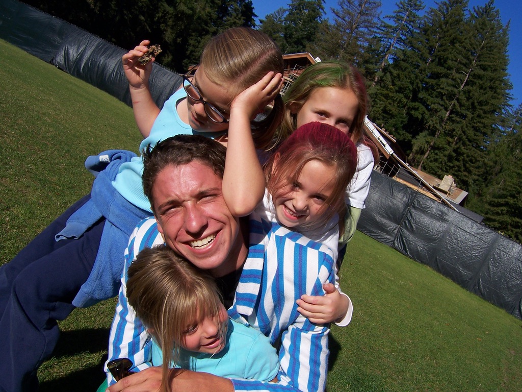 Young Ross lying on the grass at Mount Hermon with four kids piled on top of him, everyone laughing, redwoods in the background