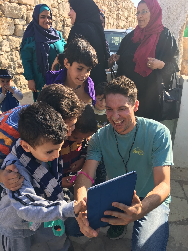 Ross kneeling with refugee children crowded around his iPad, all laughing